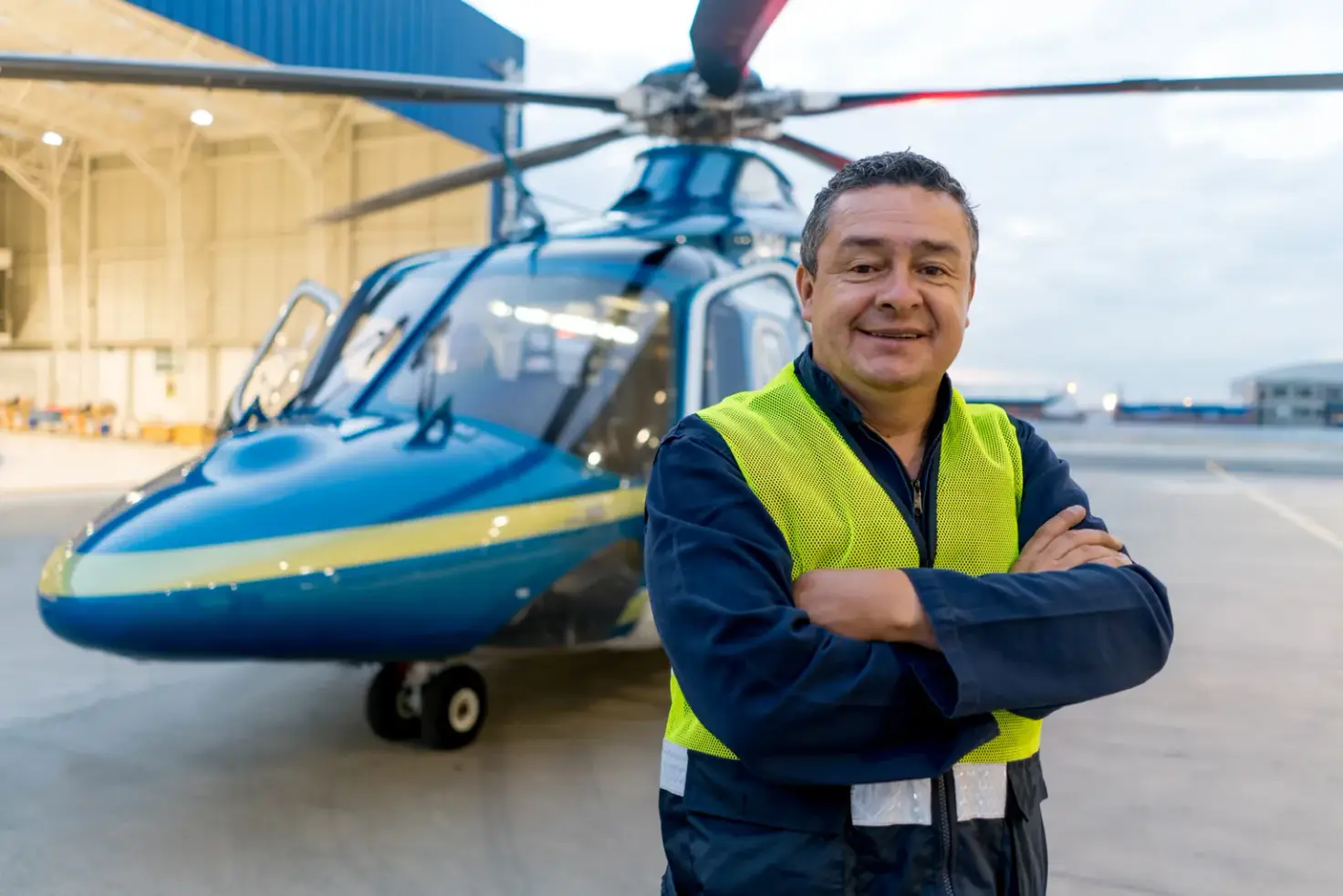 A man in a high-visibility vest stands in front of a wheeled helicopter beside an open hangar.