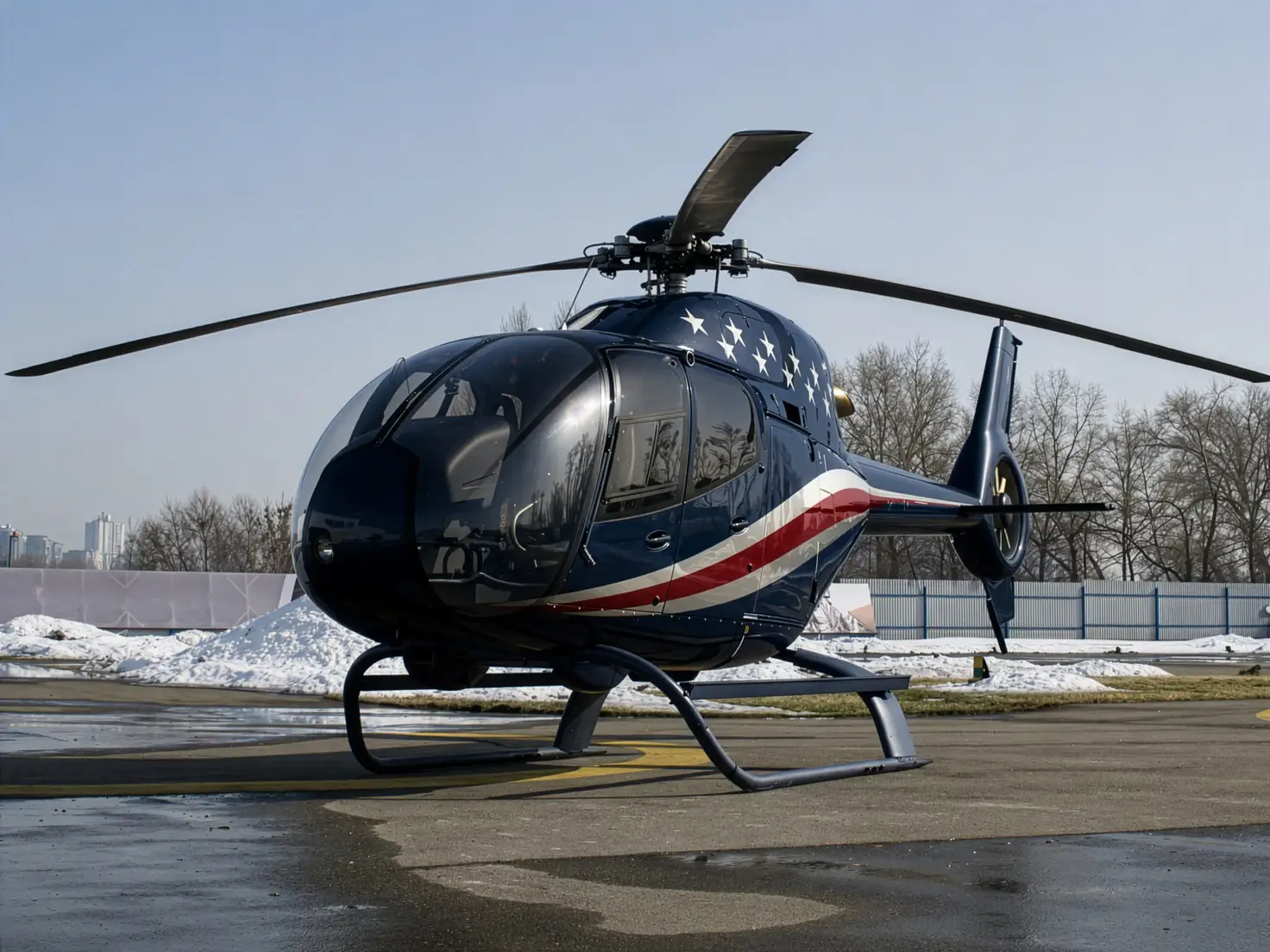 A light helicopter on skids stands on an airport apron bordered by snowbanks, with bare trees behind it.