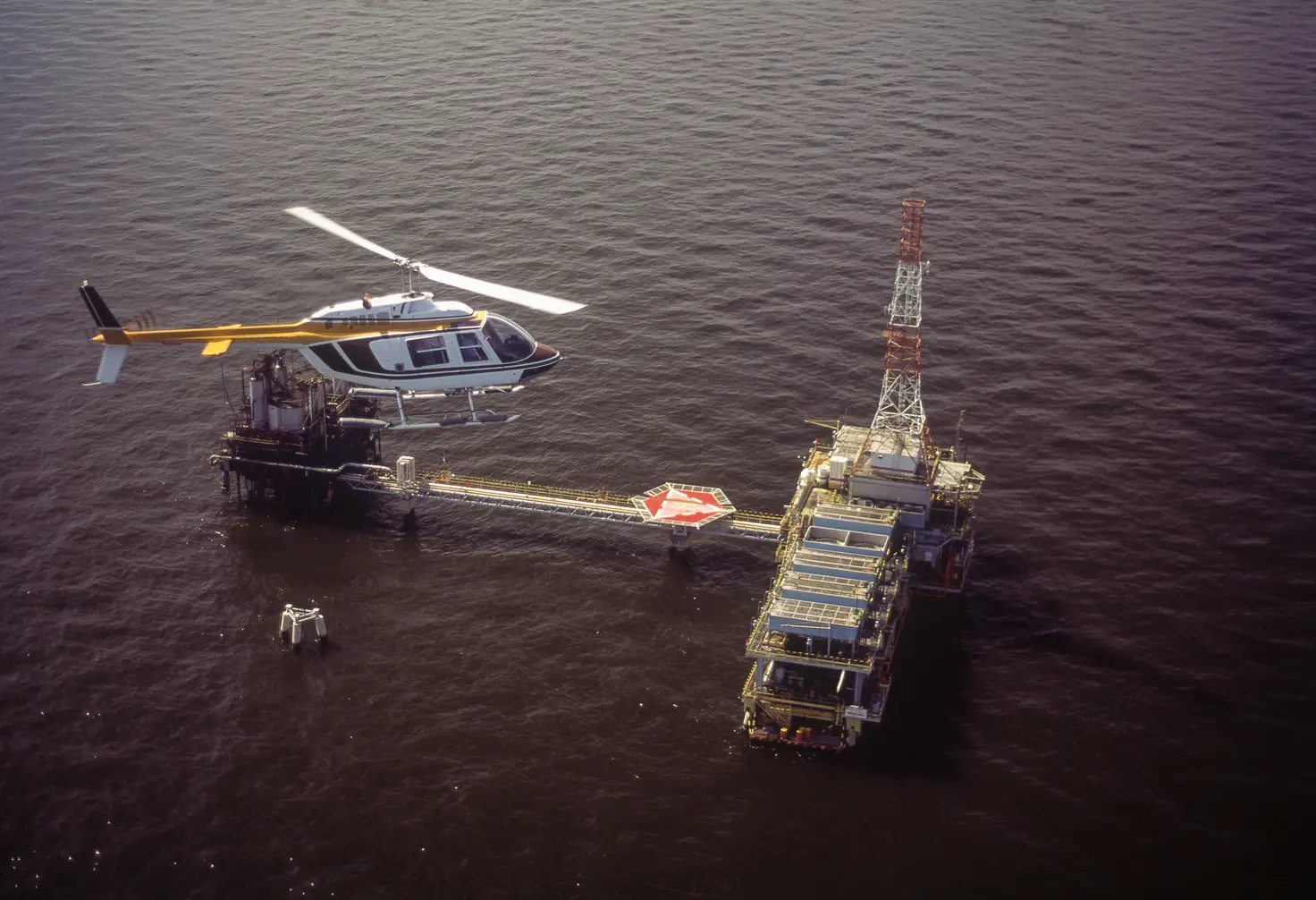 A helicopter on skids flies above the sea beside an offshore platform and its landing pad.