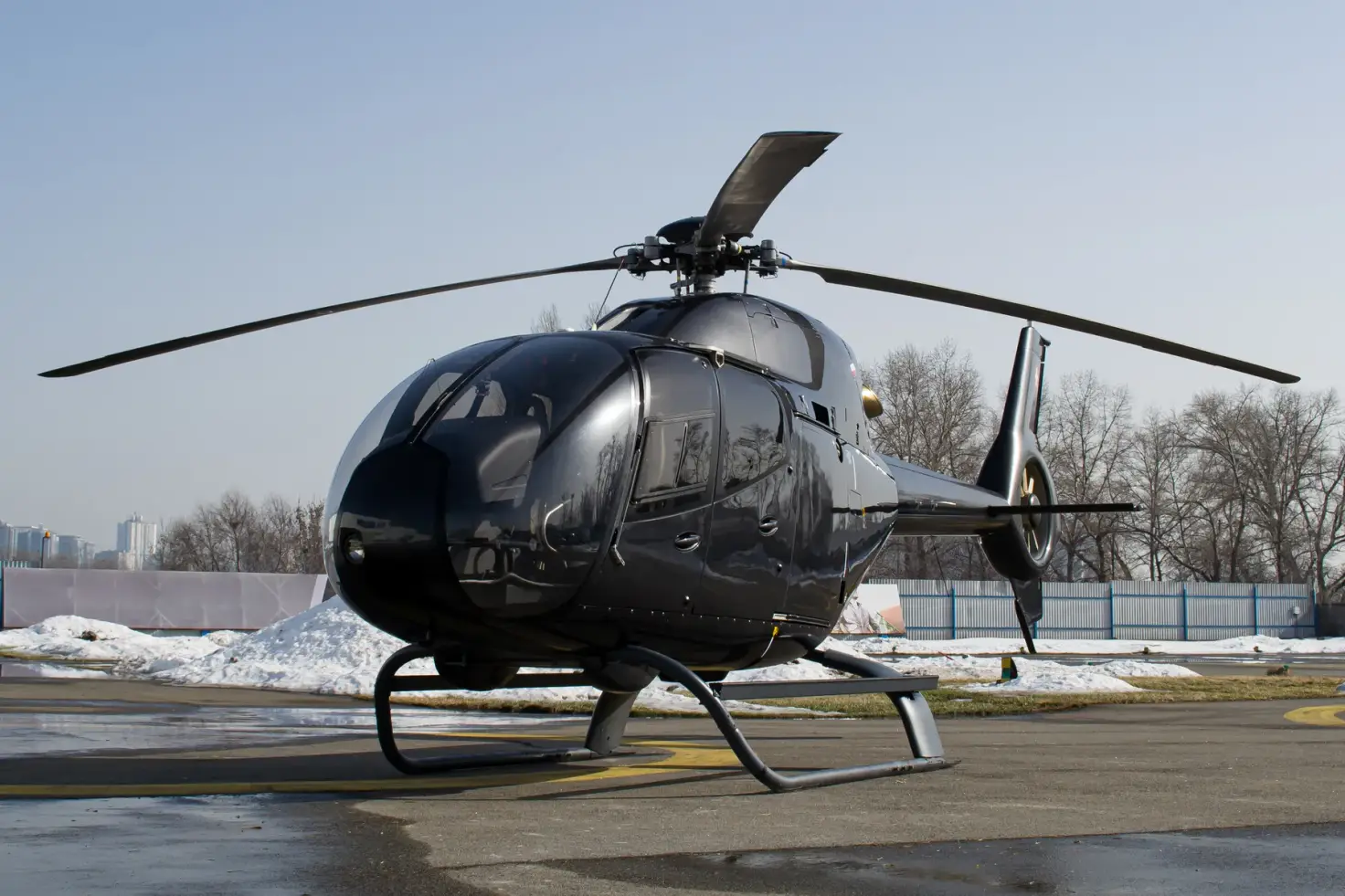 A dark light single-engine helicopter parked and shut down on a winter apron, snow banked along the fence and bare trees behind.
