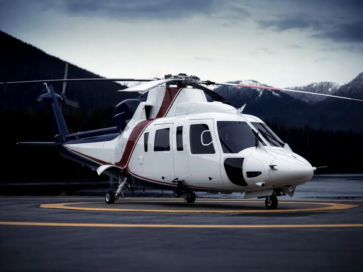 A wheeled helicopter stands on a marked landing pad with wooded mountains behind it.