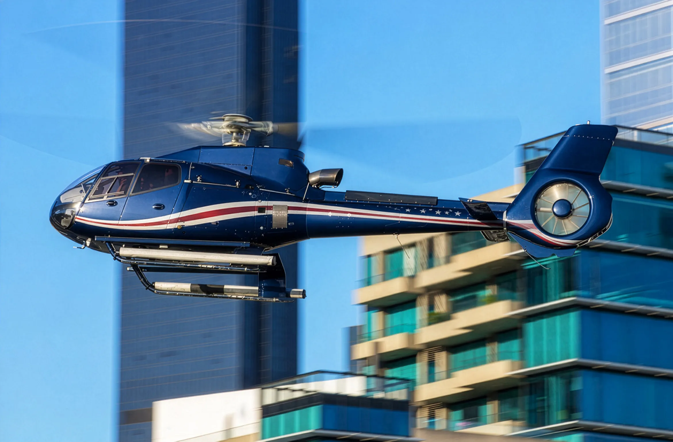 A light single-engine helicopter in low flight past city high-rise buildings.