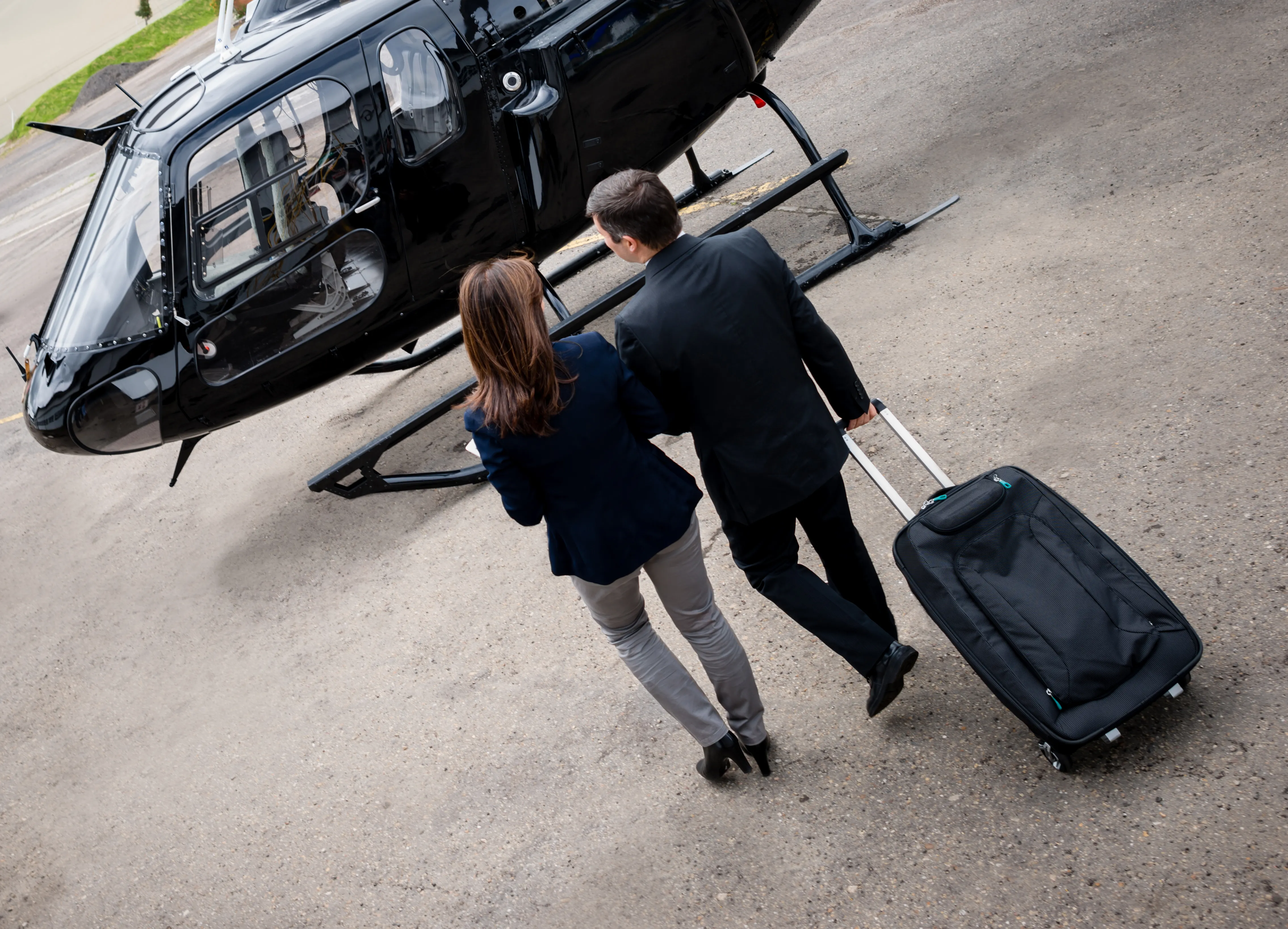 A dark light helicopter parked on an airport apron while two travelers with rolling luggage walk up to board.