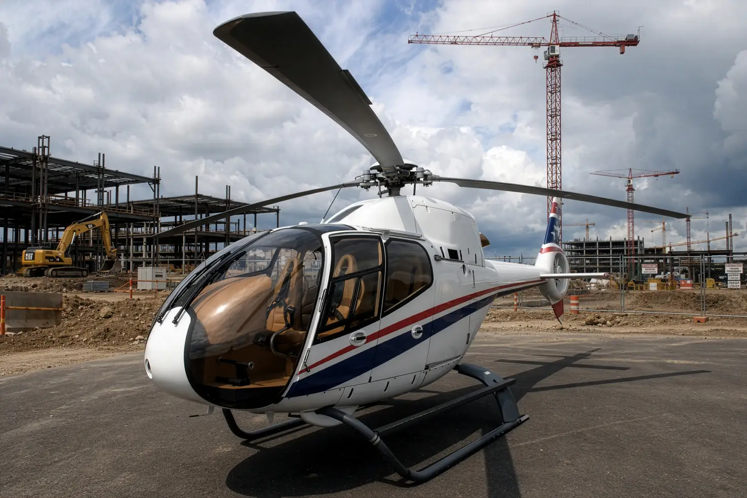 A white light single-engine helicopter with red and blue striping parked on a gravel pad at an active construction site, with tower cranes and steel framing behind it.