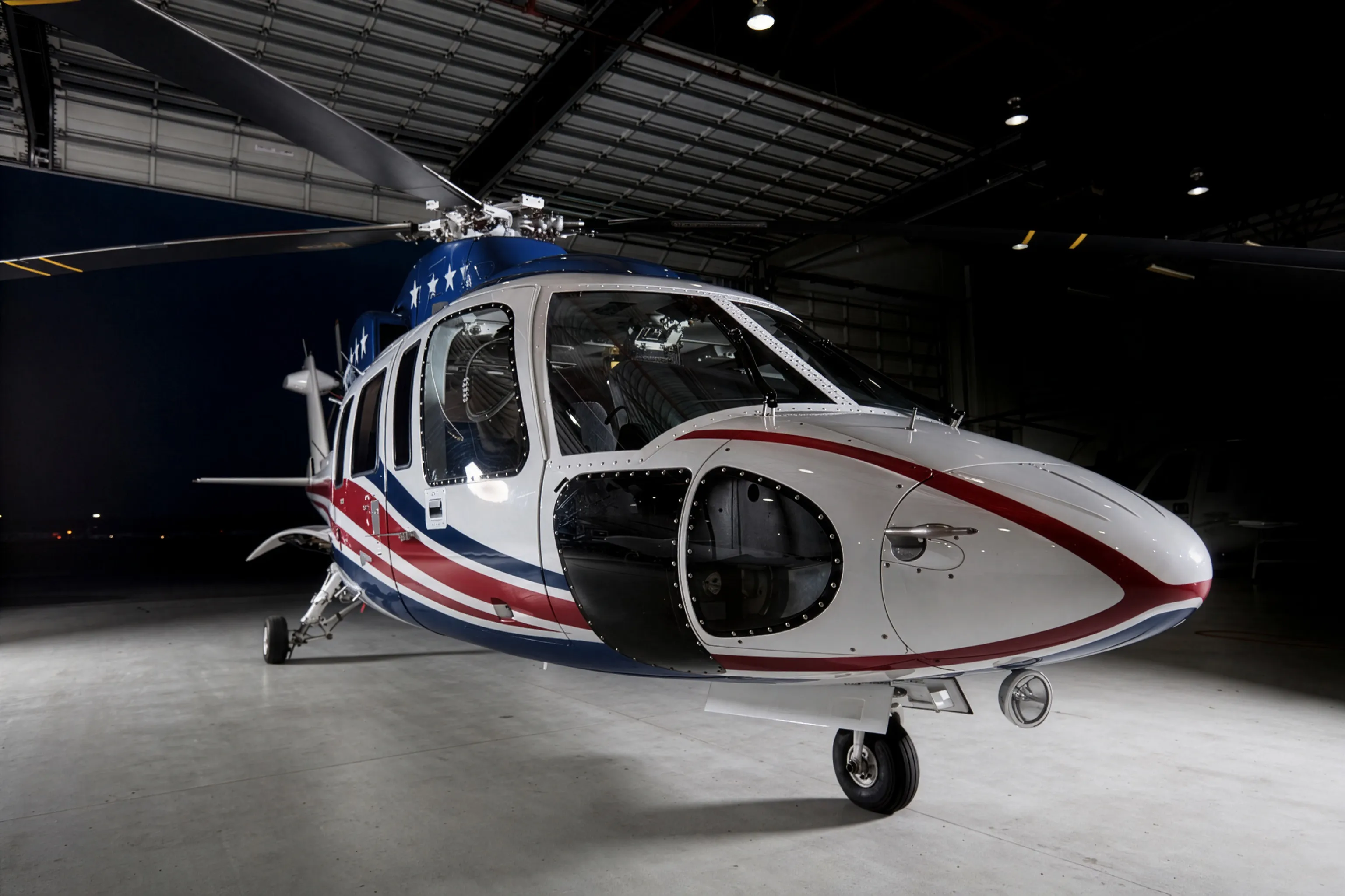 A white twin-engine executive helicopter with red and blue striping parked inside a dimly lit hangar, staged ahead of an early, low-profile departure.