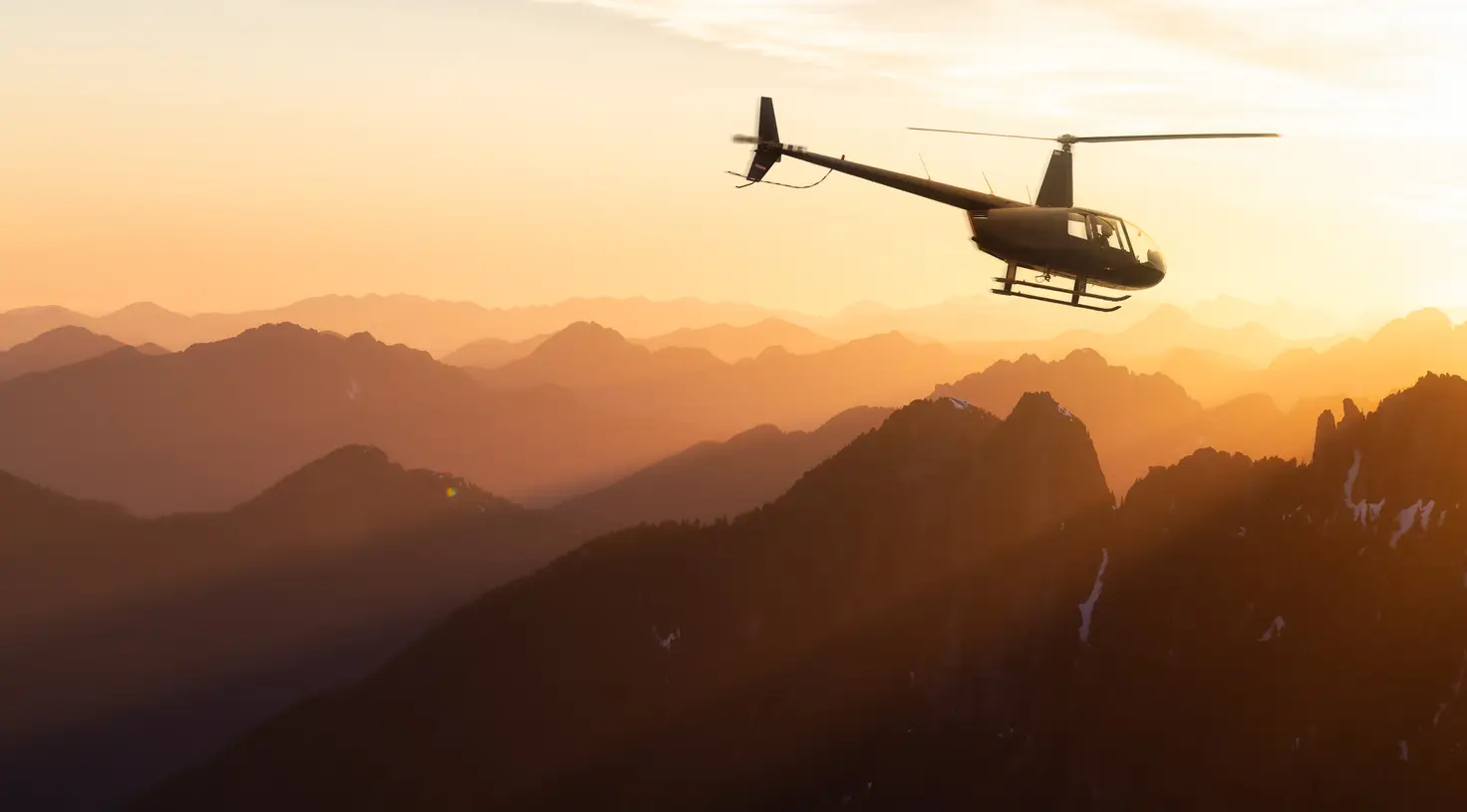 A light single-engine helicopter silhouetted against a golden sunset, flying above layered mountain ridges on a scenic charter.