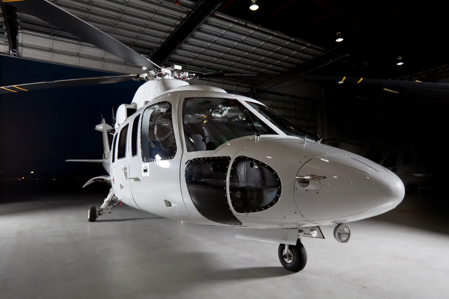 A white helicopter on wheeled landing gear stands inside a lit hangar at night.