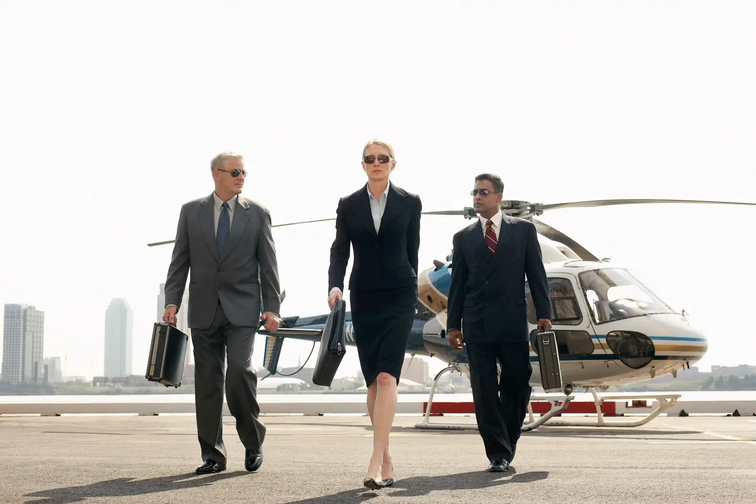 Three travelers with briefcases walking away from a light helicopter parked on a rooftop helipad with a city skyline behind.