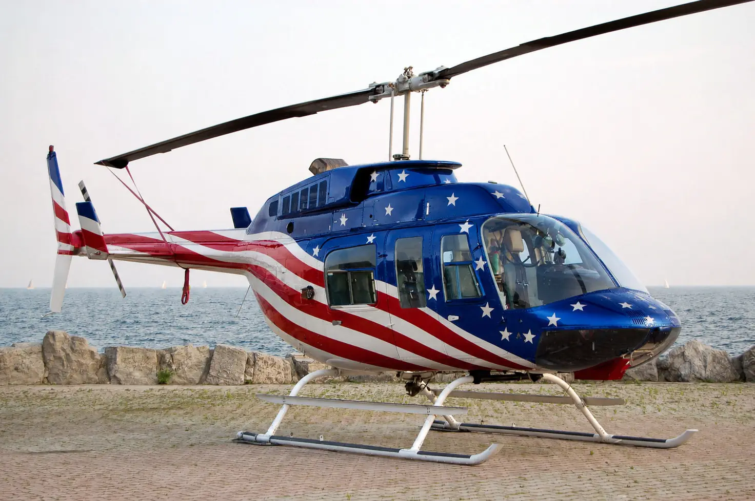 A helicopter on skids is parked beside a seawall, with open water and sailboats behind it.