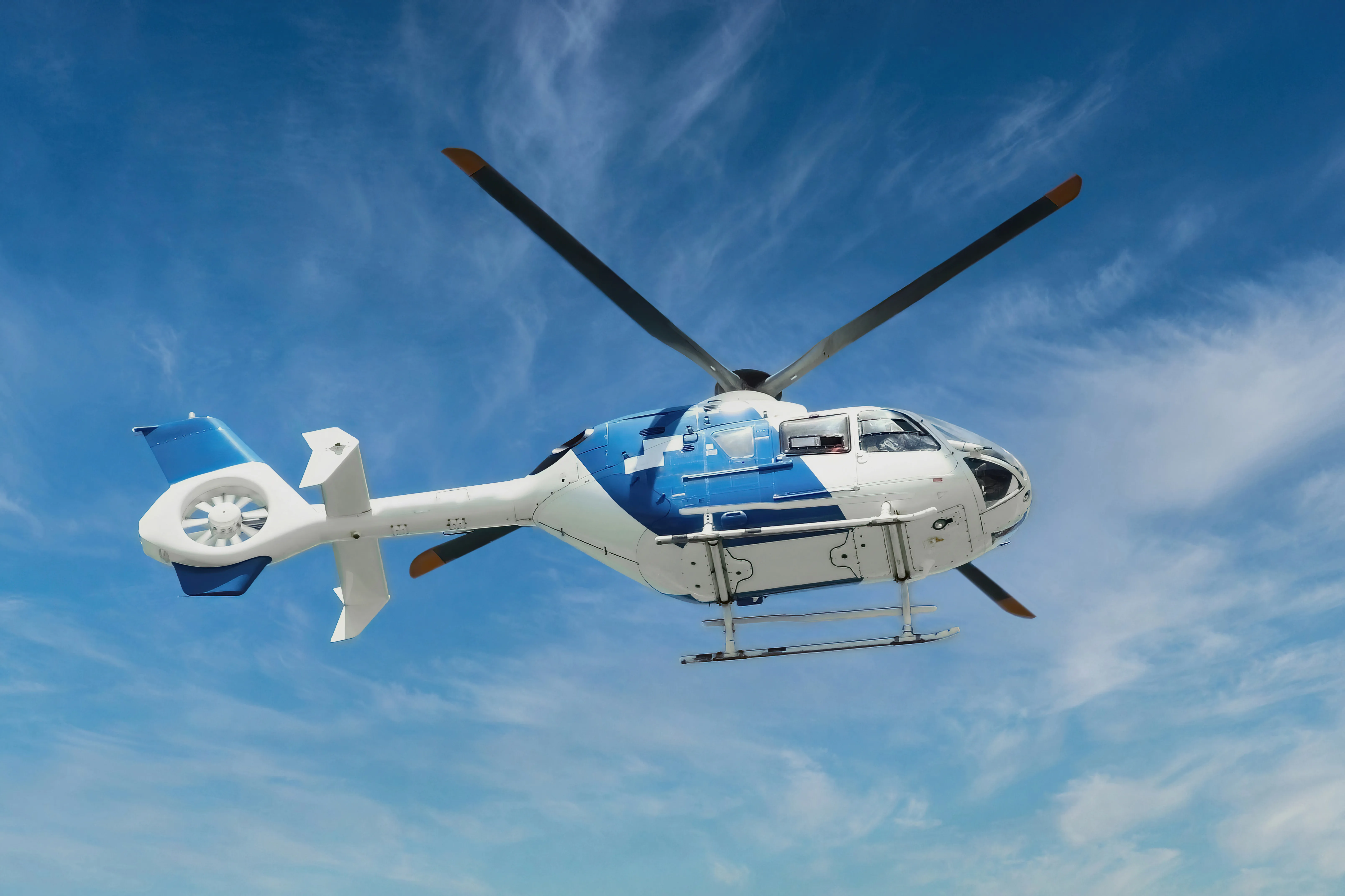 A white and blue light twin-engine helicopter in flight against a bright sky, the view from the drive-versus-flight trade a weekend charter makes.