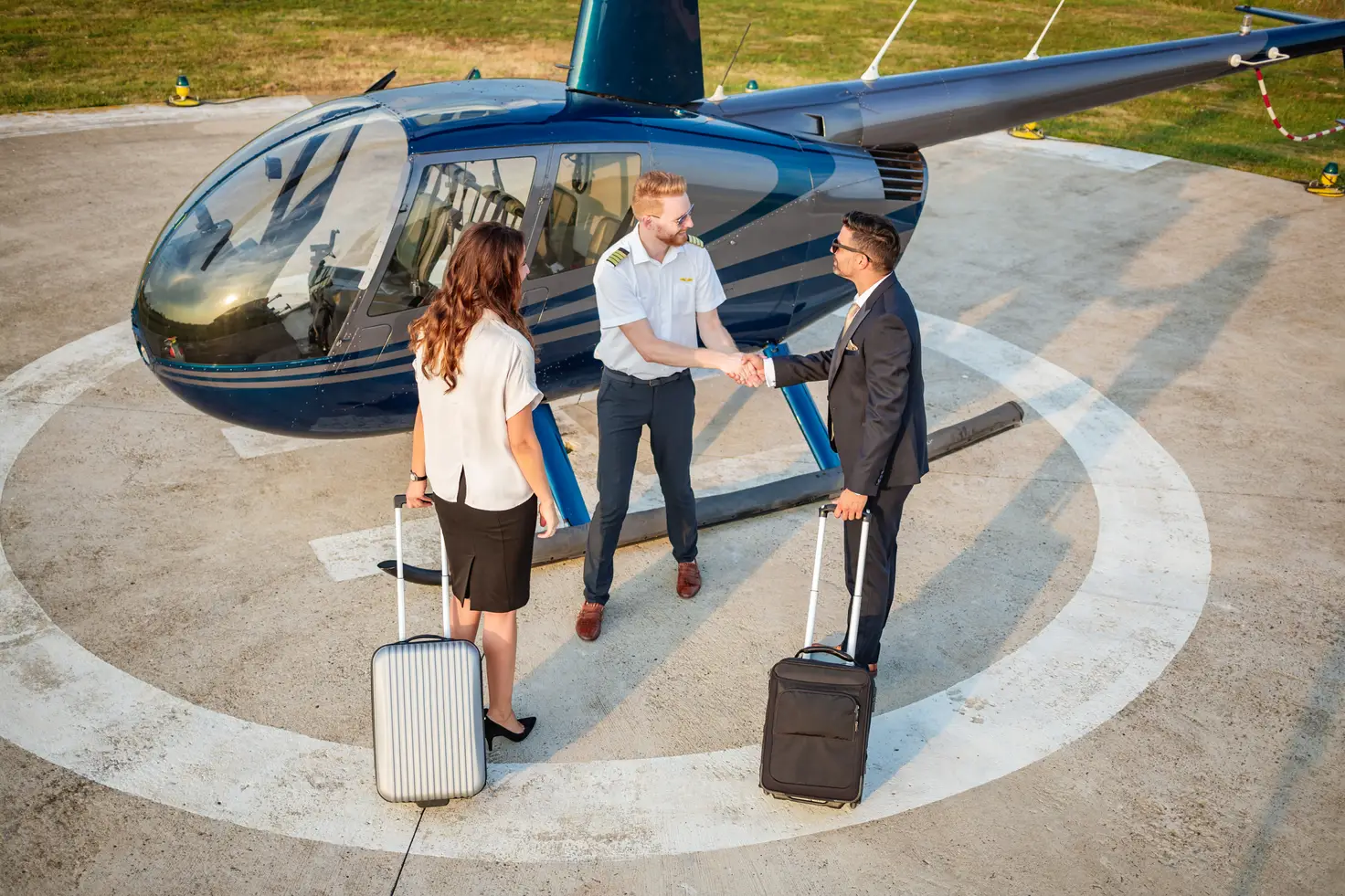 A pilot greeting two passengers with rolling luggage beside a dark blue light helicopter parked on a marked helipad.