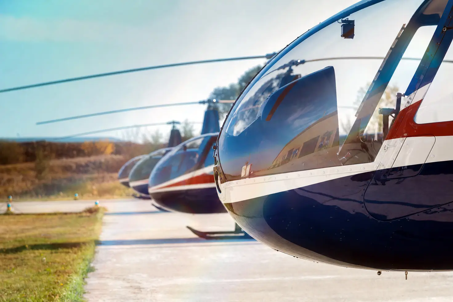 A row of light single-engine helicopters parked nose to tail on a sunny airport flight line, the class most often used for sightseeing charters.