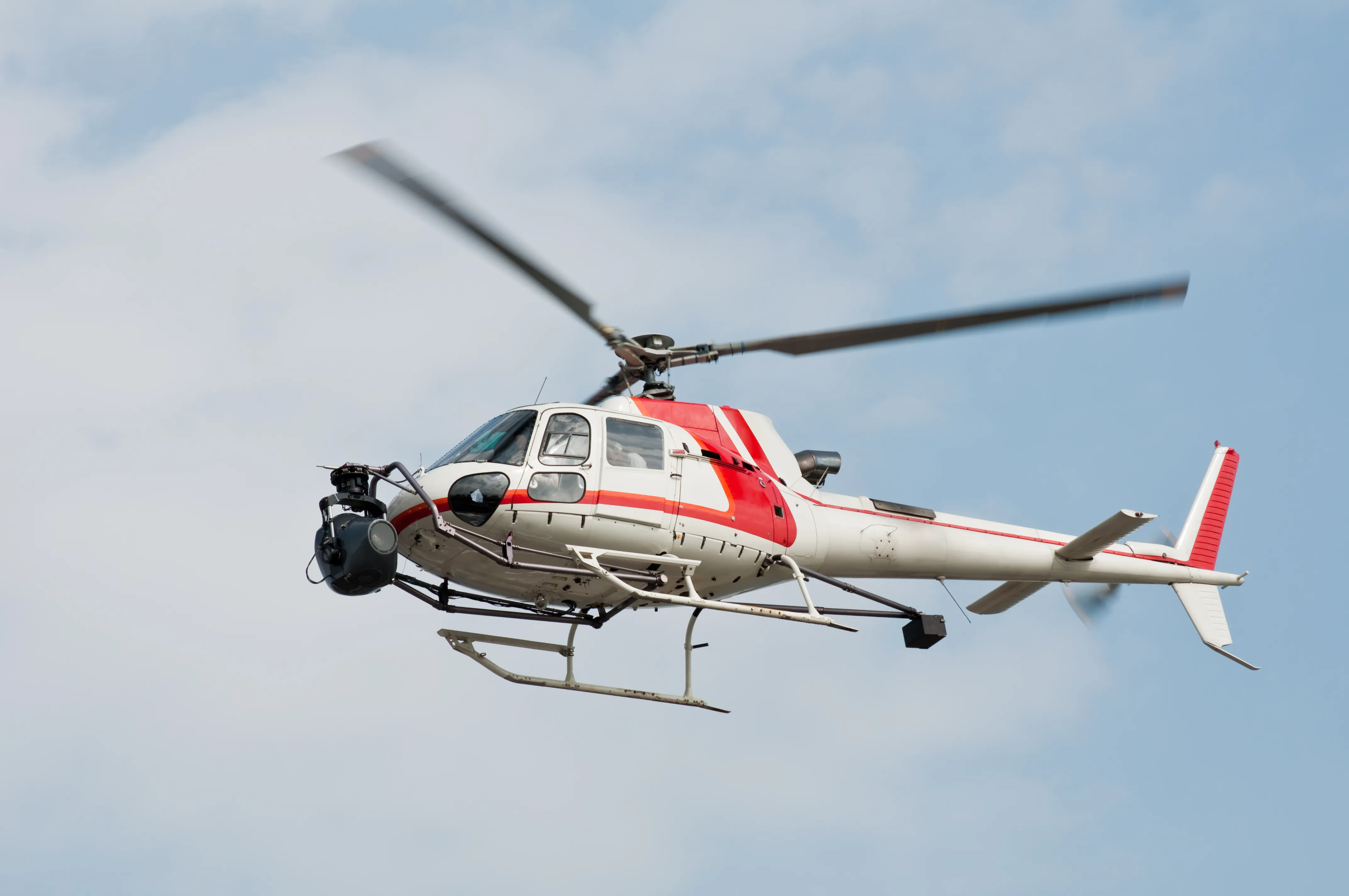 A white and red light single-engine helicopter in flight carrying a gyro-stabilized camera gimbal mounted beneath its nose.
