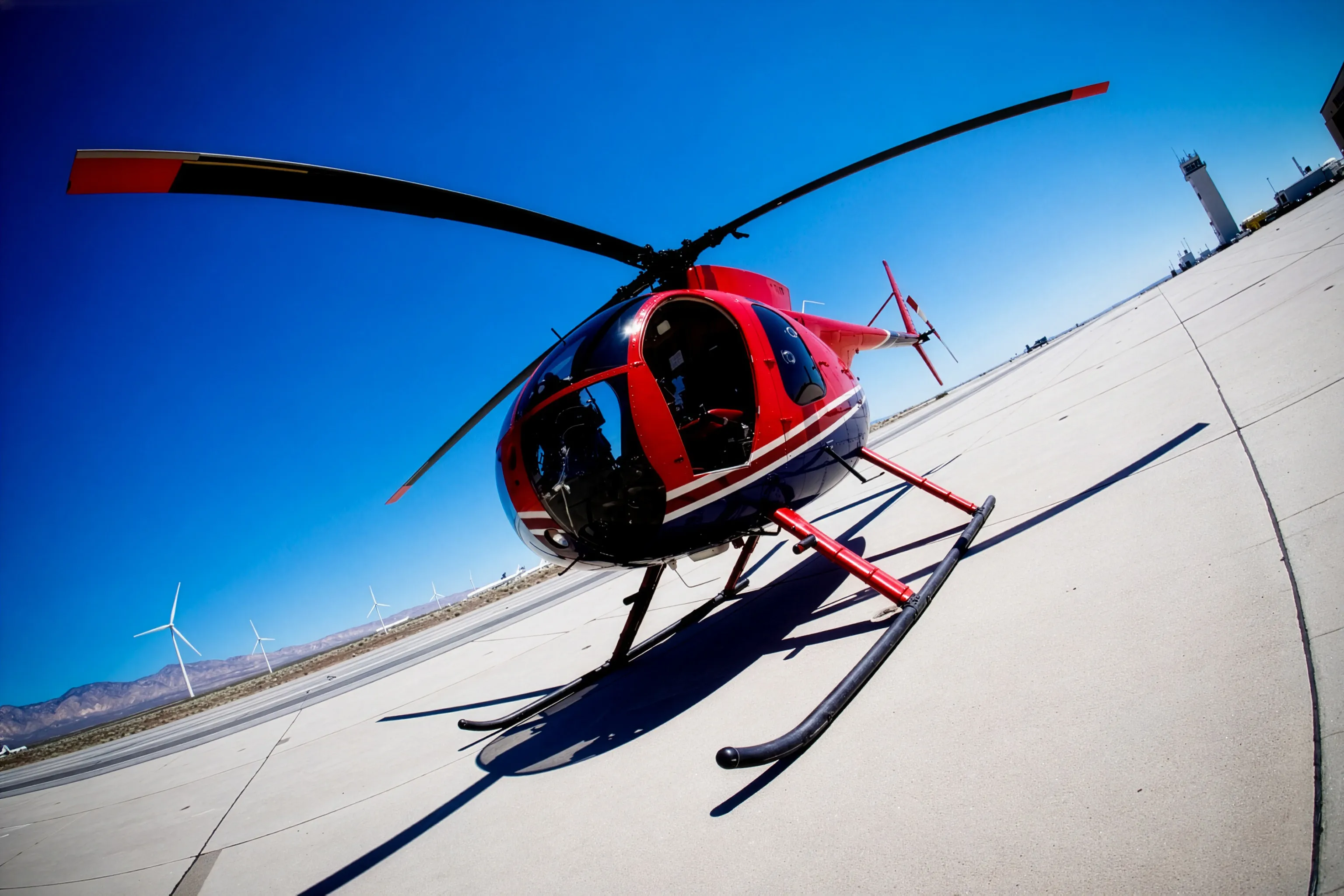 A red, white, and blue MD 500 light helicopter with skid landing gear parked on a desert airfield apron, wind turbines on the horizon behind it.