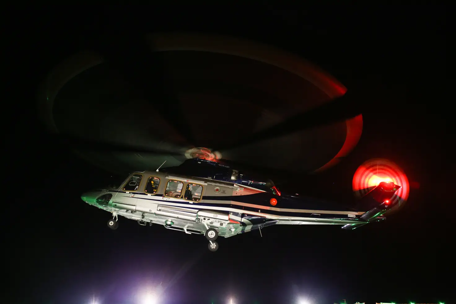 A helicopter lifts above a floodlit landing area at night with its landing gear extended.