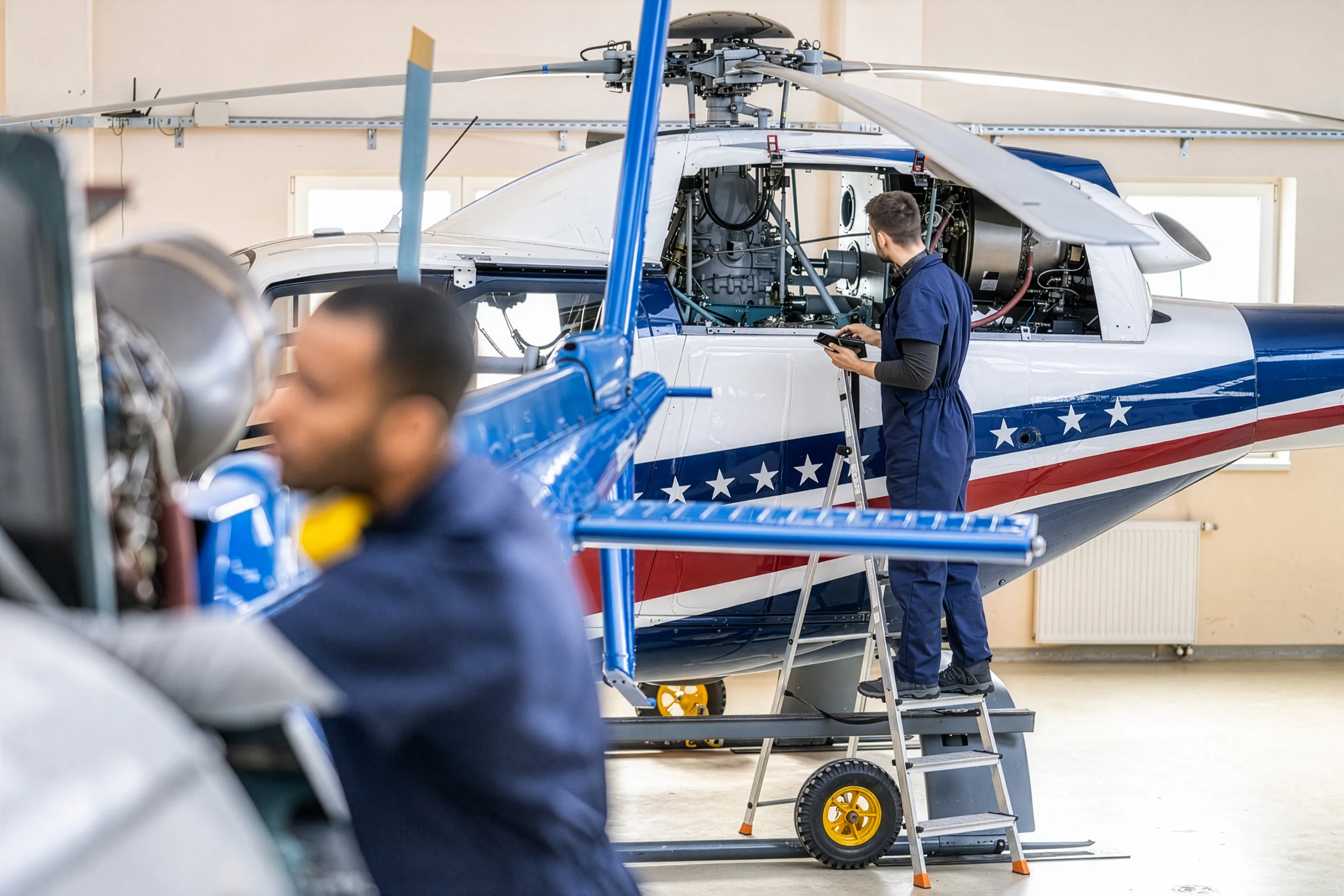 A helicopter in a maintenance hangar with its engine compartment open and a technician on a ladder, the kind of grounded aircraft an AOG parts charter serves.